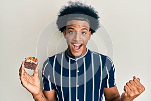 African american man with afro hair holding cake slice screaming proud, celebrating victory and success very excited with raised