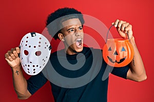 African american man with afro hair wearing hockey mask and halloween pumpking angry and mad screaming frustrated and furious,