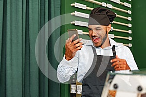 African american man barista using his smartphone and laughing while standing at the counter of coffee shop