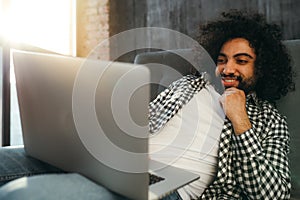 African american man with a black beard in a white t-shirt sits on the couch looking at the computer