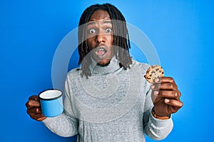 African american man with braids drinking a cup of coffee and cookie afraid and shocked with surprise and amazed expression, fear