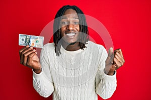 African american man with braids holding 100 dollars banknote screaming proud, celebrating victory and success very excited with