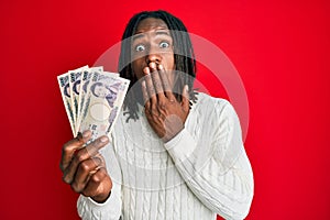 African american man with braids holding 5000 japanese yen banknotes covering mouth with hand, shocked and afraid for mistake