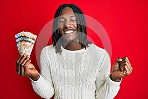 African american man with braids holding canadian dollars screaming proud, celebrating victory and success very excited with