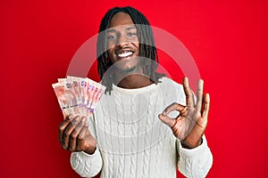 African american man with braids holding south african 50 rand banknotes doing ok sign with fingers, smiling friendly gesturing