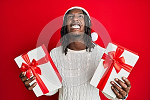 African american man with braids wearing christmas hat and holding gifts angry and mad screaming frustrated and furious, shouting