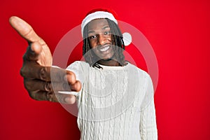 African american man with braids wearing christmas hat smiling friendly offering handshake as greeting and welcoming