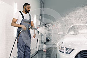 African American man, car wash worker is spraying cleaning foam to a modern white luxury car holding a high pressure