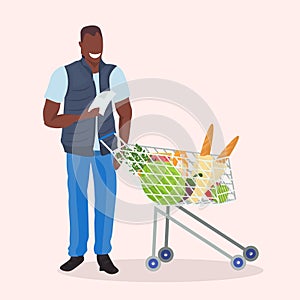 African american man checking shopping list happy male customer with trolley cart buying products in grocery market