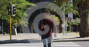 African american man in city, using smartphone, wearing headphones and backpack crossing street