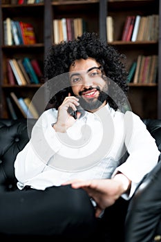 An African-American man with curly hair is talking on the phone, sitting on the sofa in the library