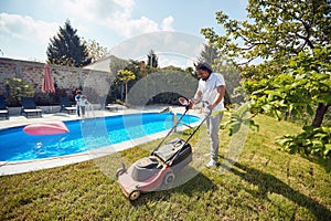 African american man cutting grass with electric grass mower by the pool, with his kids playing by the pool.African american man