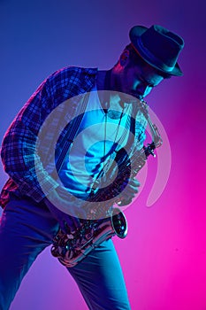 African-American man dressed retro, in hat playing saxophone against gradient pink and blue background in neon light.