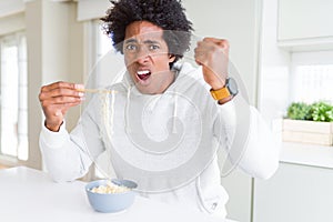 African American man eating asian noodles using chopsticks at home annoyed and frustrated shouting with anger, crazy and yelling