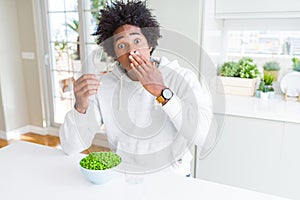 African American man eating fresh green peas at home cover mouth with hand shocked with shame for mistake, expression of fear,