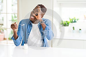 African american man eating healthy natural yogurt with a spoon with happy face smiling doing ok sign with hand on eye looking