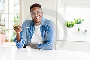 African american man eating healthy natural yogurt with a spoon with a happy face standing and smiling with a confident smile