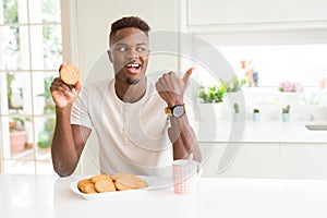 African american man eating healthy whole grain biscuit pointing and showing with thumb up to the side with happy face smiling