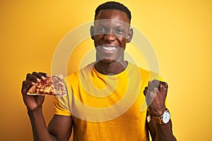 African american man eating slice of pizza standing over isolated yellow background screaming proud and celebrating victory and
