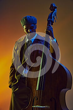 African-American man, elegant bassist in trendy hat engaged in music in neon light against gradient background.