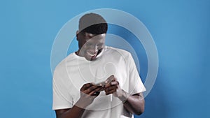 an African-American man gambles in a smartphone while standing on a blue background.