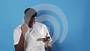 an African-American man gambles in a smartphone while standing on a blue background.