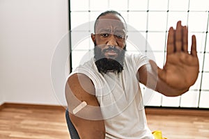 African american man getting vaccine showing arm with band aid with open hand doing stop sign with serious and confident
