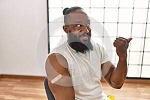 African american man getting vaccine showing arm with band aid pointing thumb up to the side smiling happy with open mouth