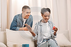 african american man gifting ring in heart shaped box to woman sitting on couch