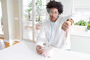 African American man holding stethoscope checking financial health on piggy bank with angry face, negative sign showing dislike