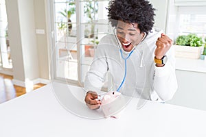African American man holding stethoscope checking financial health on piggy bank screaming proud and celebrating victory and