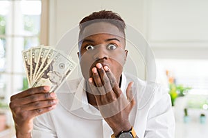 African american man holding twenty dollars bank notes cover mouth with hand shocked with shame for mistake, expression of fear,