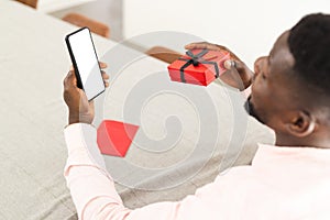 African American man holds a phone with a blank screen and a small red gift box with copy space