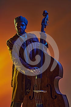 African-American man, jazz musician with beard, bowing cello instrument in neon light against gradient background.