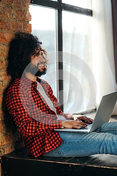 African American man in jeans sits on the floor against the background of a window at a computer