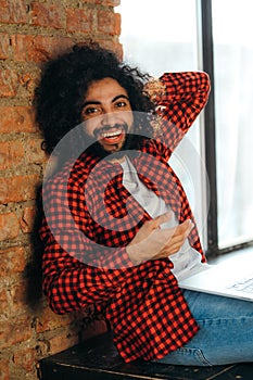 African American man in jeans sits on the floor against the background of a window at a computer