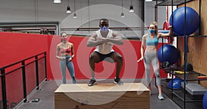 African american man jumping on box wearing face mask at gym, two diverse women watching