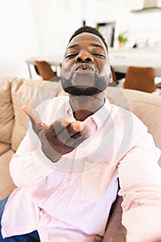 African American man in a pink shirt blows a kiss on video call, seated on a beige sofa