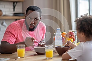 African american man in a pink tshirt feeding his kid and frowing to entertain her photo