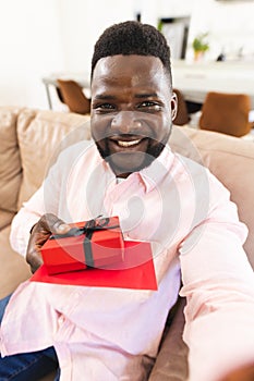 African American man presents a red gift box with a smile, sitting indoors on video call