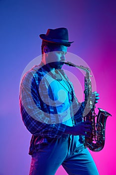 African-American man, saxophonist dressed retro, in hat playing against gradient pink and blue background in neon light.