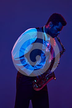 African-American man in shirt and vest playing saxophone against blue and purple background in neon lighting. Jazz and