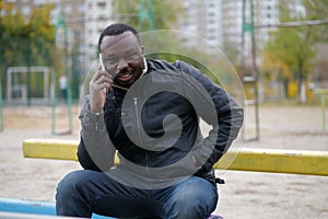 African american man sits at sport ground and speaks on smartphone