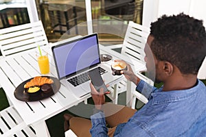 African american man sitting in cafe using laptop and smartphone drinking coffee eating breakfast