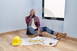 African american man sitting on the floor at new home looking at blueprints covering one eye with hand, confident smile on face