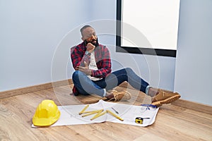 African american man sitting on the floor at new home looking at blueprints with hand on chin thinking about question, pensive