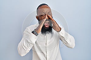 African american man standing over blue background shouting angry out loud with hands over mouth