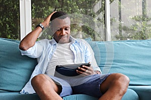 African American man using digital tablet while sitting on cozy sofa couch at home