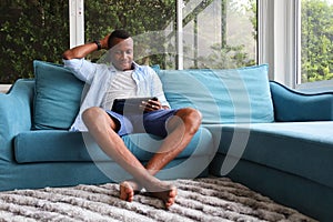 African American man using digital tablet while sitting on cozy sofa couch at home