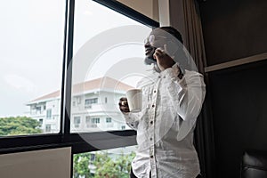 African American man using have call by phone while drink coffee by the window in the living room